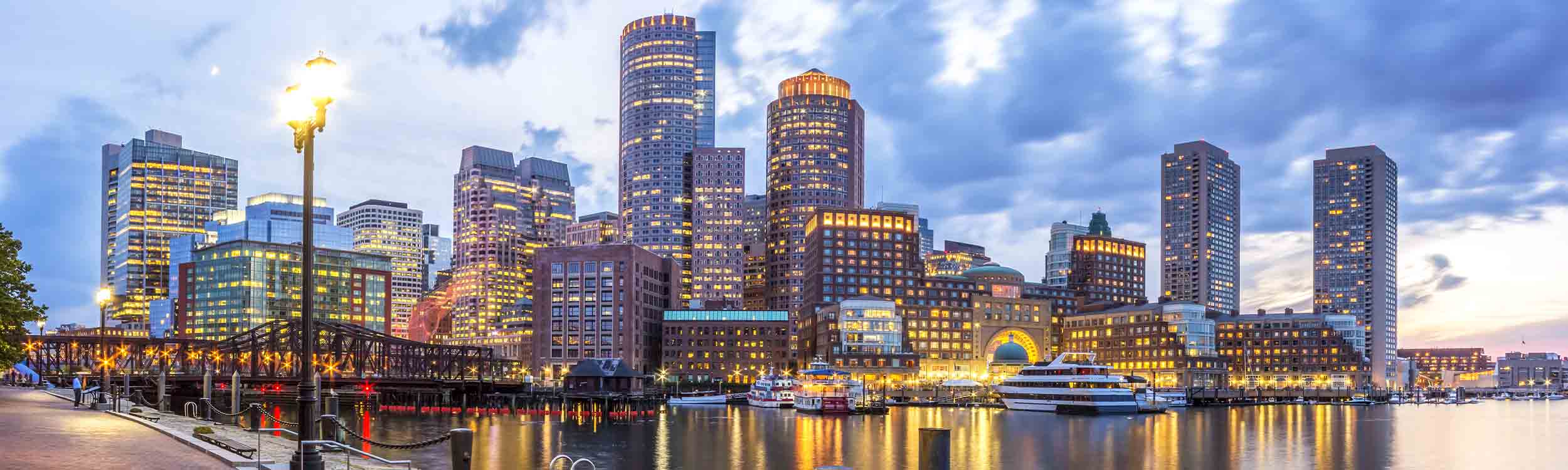 Panoramic view of the Boston skyline and Harbor at twilight, featuring the illuminated Rowes Wharf.